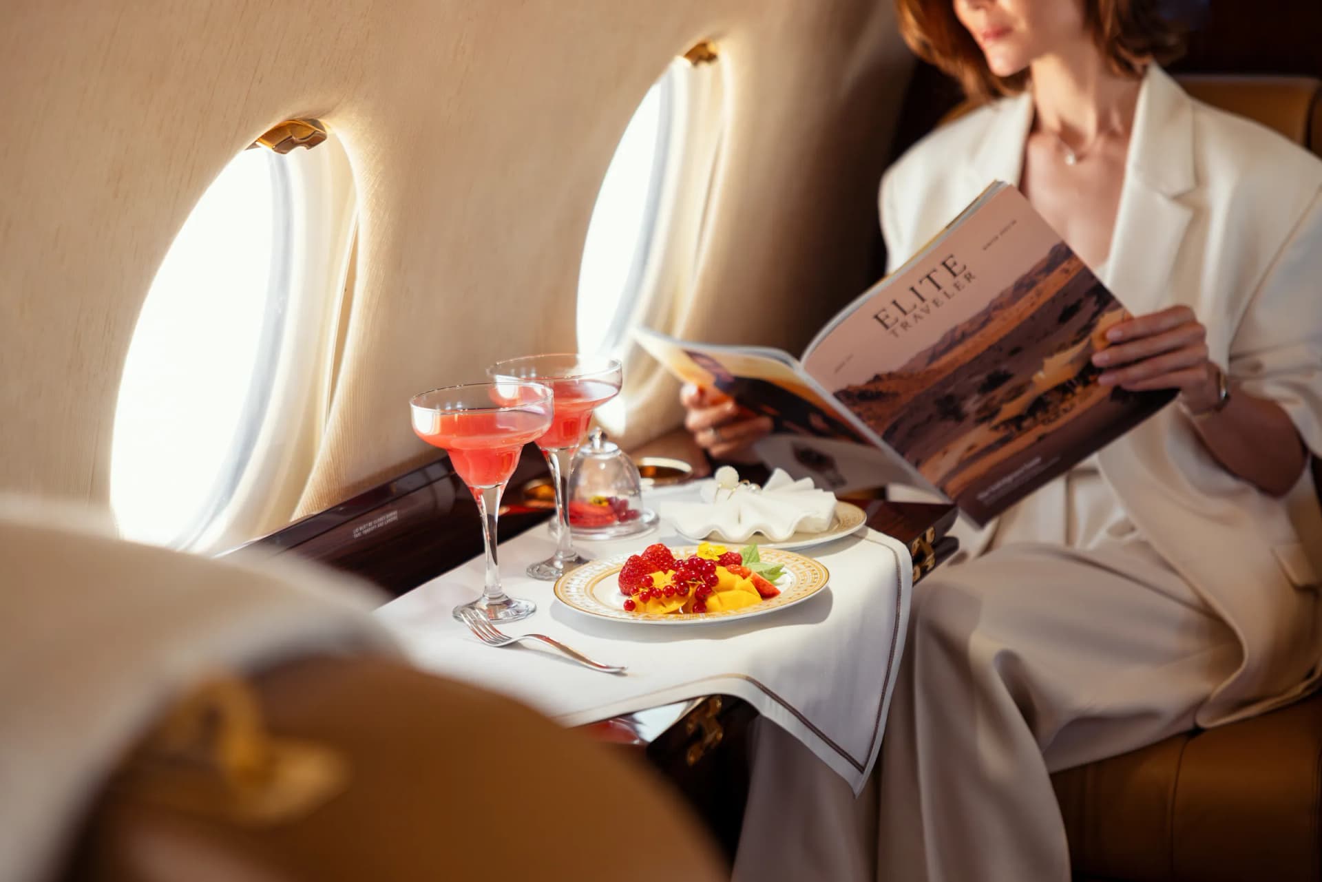 A guest reading a magazine at a table with cocktails and a fruit plate, by a Gulfstream G200 window