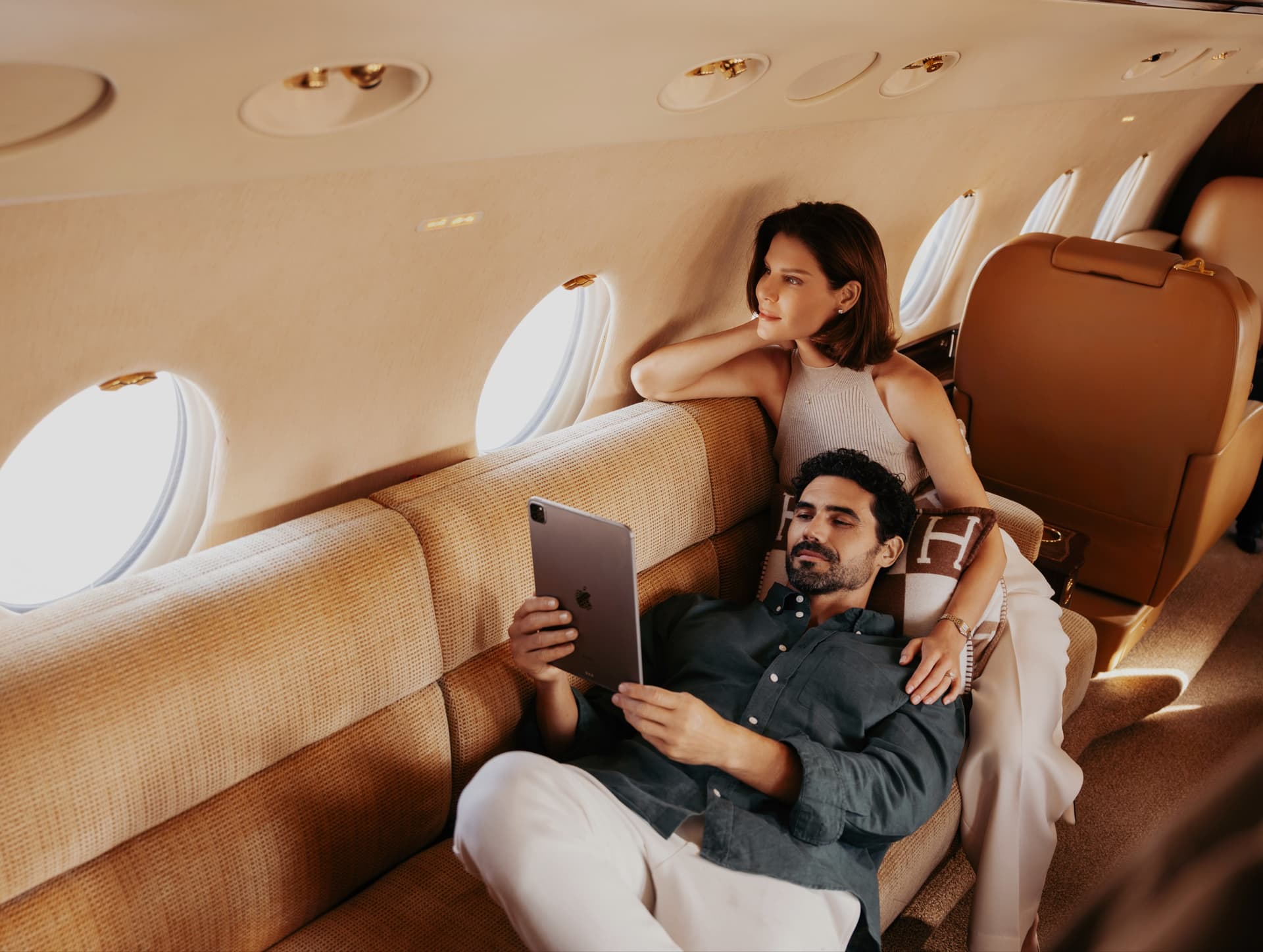 Couple relaxing in a private jet cabin — reading on a tablet beside sunlit portholes
