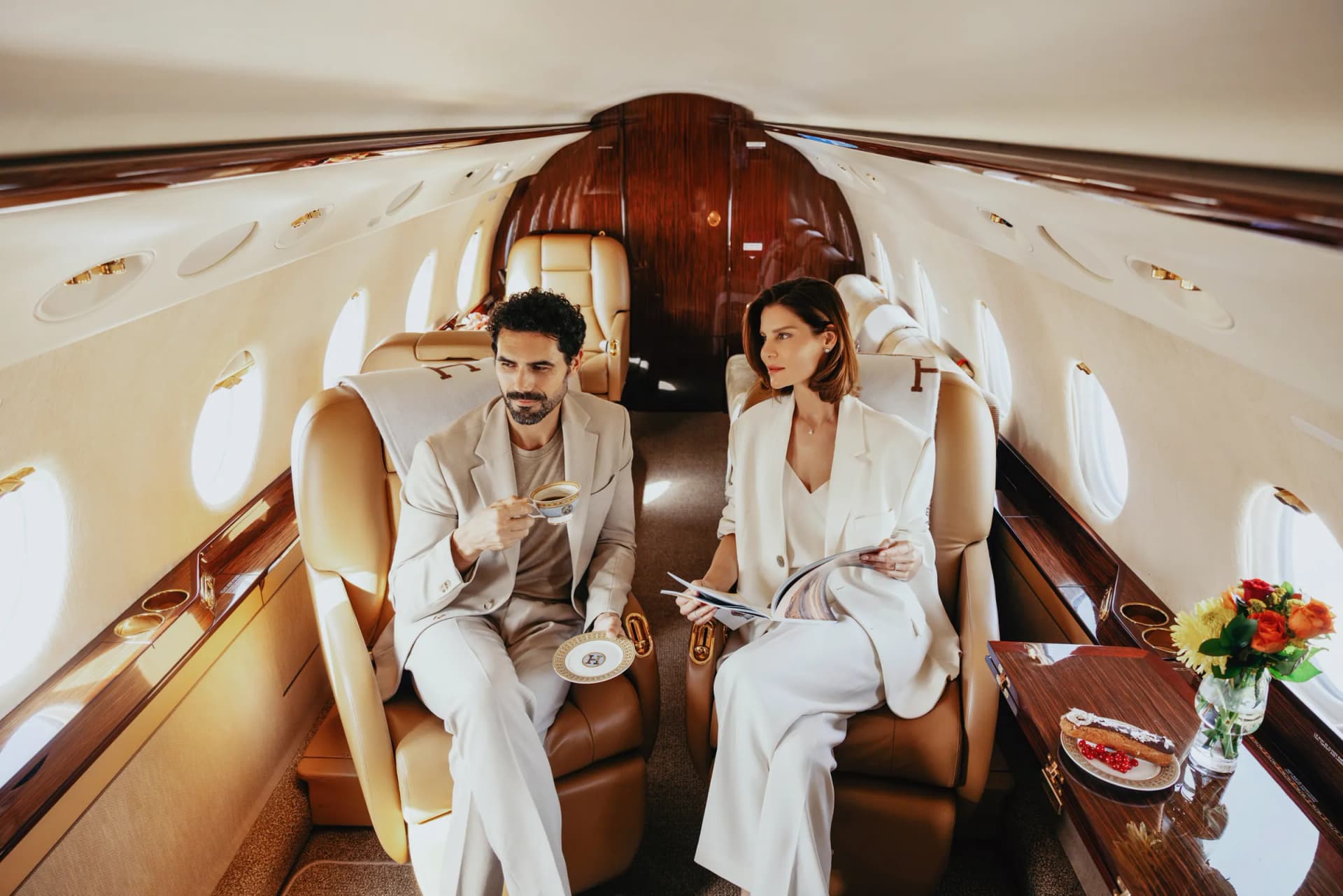 Two guests in tan leather club seats in the Gulfstream G200 cabin, with coffee and a magazine