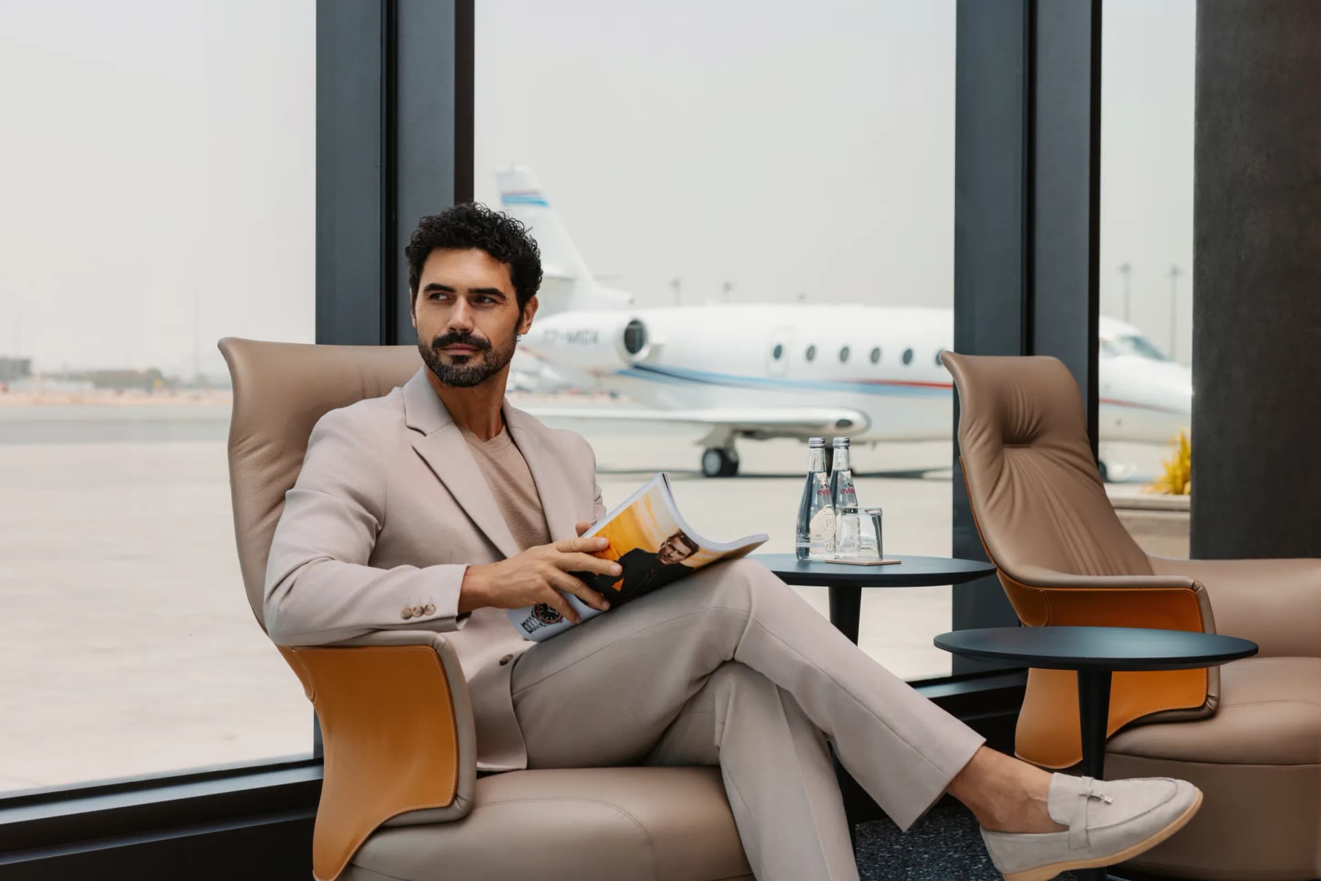 A traveller waiting in a lounge chair by the window, a private jet on the apron behind him