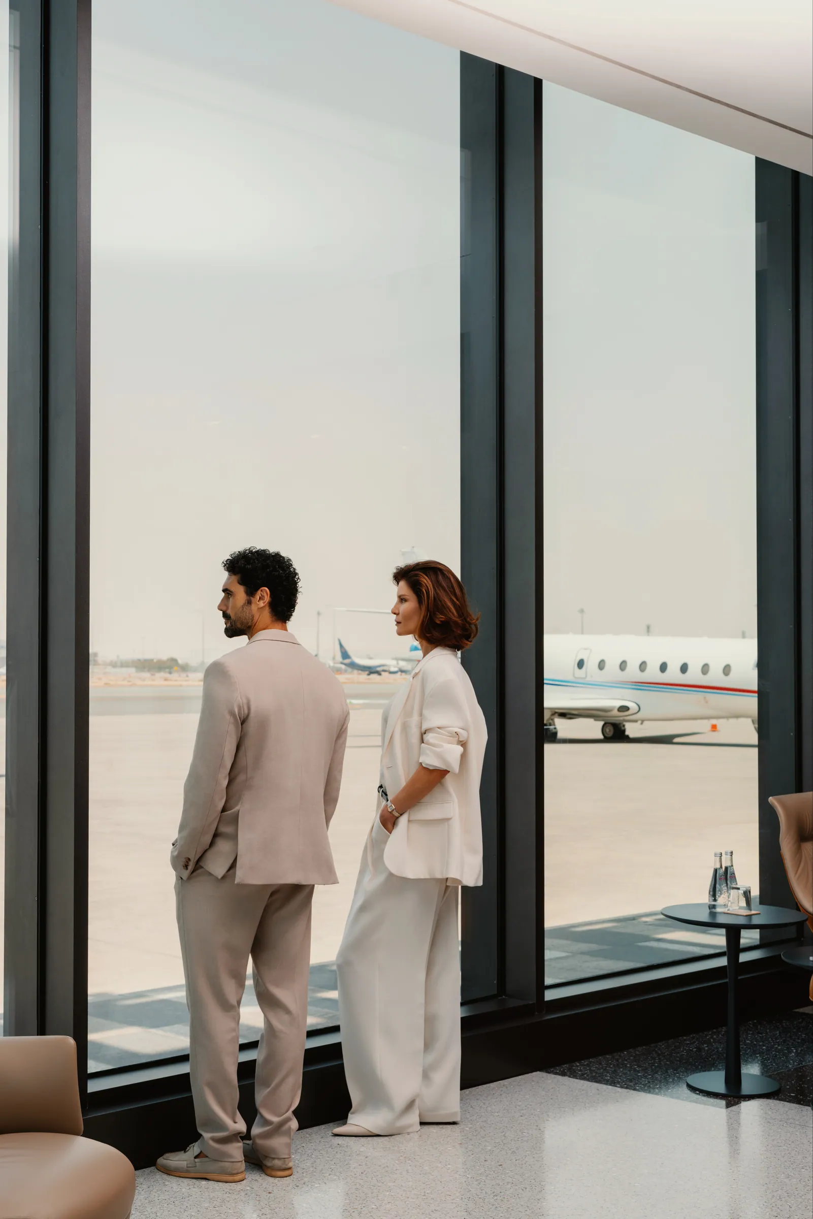 Two travellers looking out at a private jet on the apron from the lounge windows
